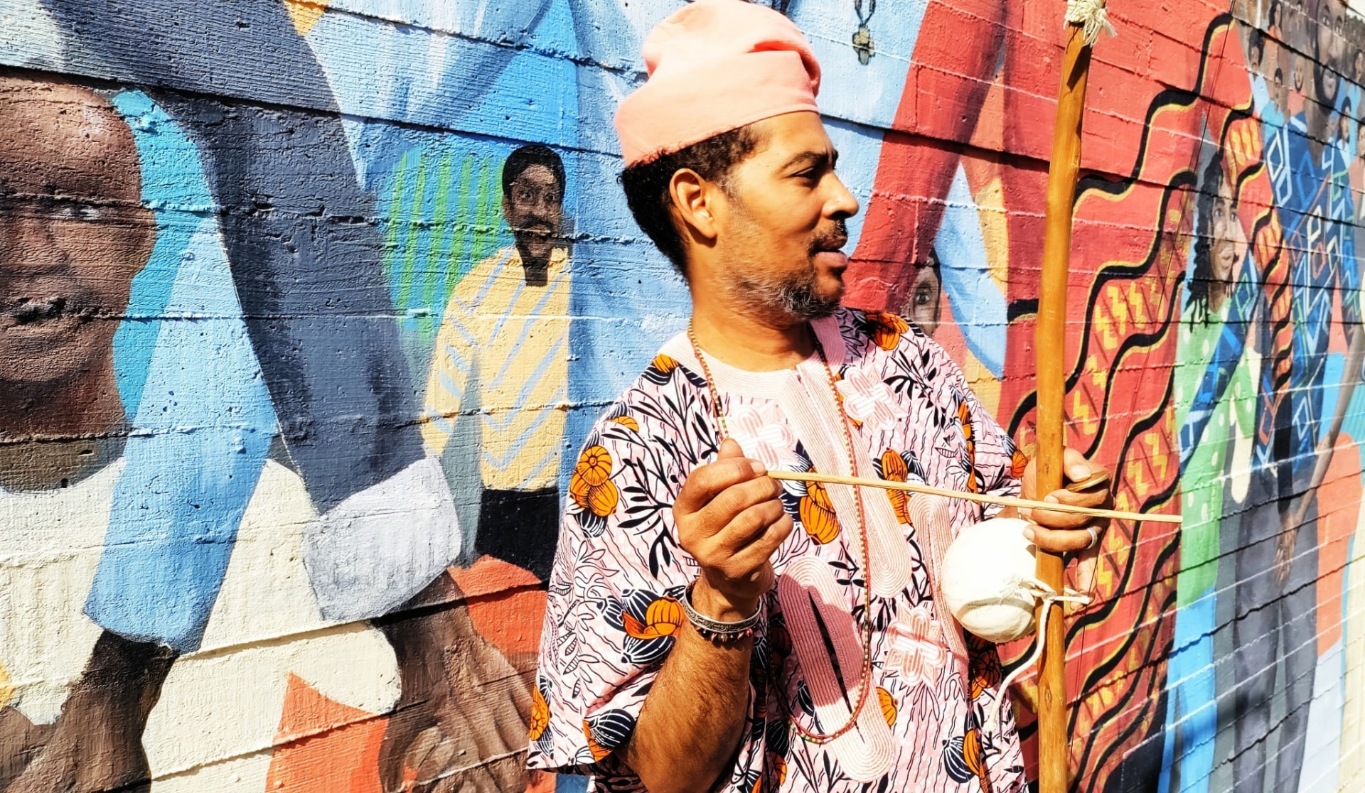 Awó, Bàbá Èṣùdámiláre playing a berimbau in front of a colorful community mural